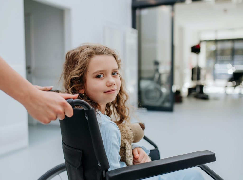 Portrait of little girl with teddy bear sitting on wheelchair.