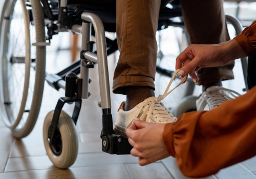 Hands of young female carer tying shoelace on footwear of man with disability